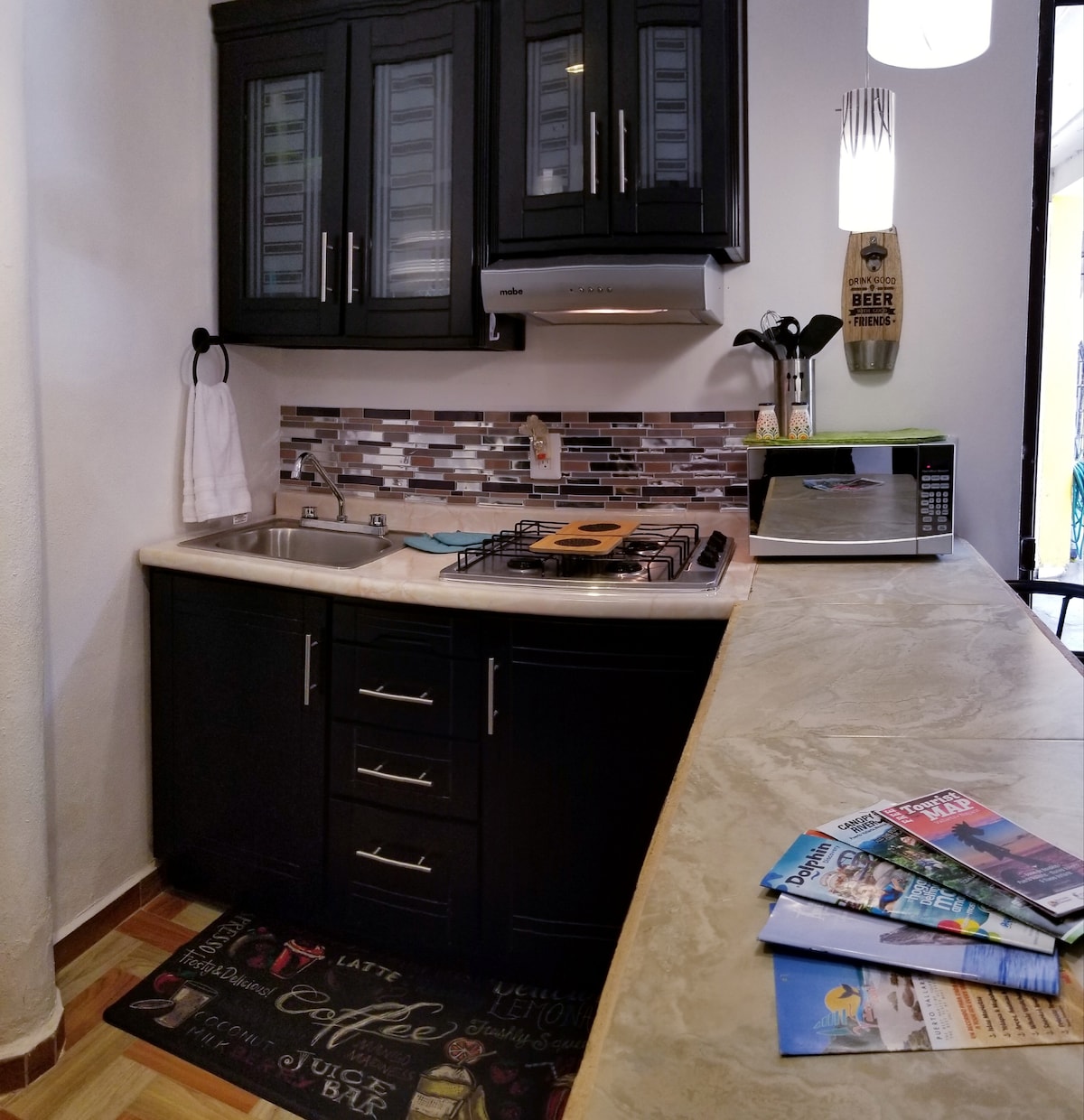 A compact kitchen area features modern black cabinetry and a countertop with a marble finish. Stainless steel appliances include a gas stove and microwave. A decorative backsplash adds a touch of style. On the countertop, various magazines are neatly displayed, contributing to the inviting atmosphere.