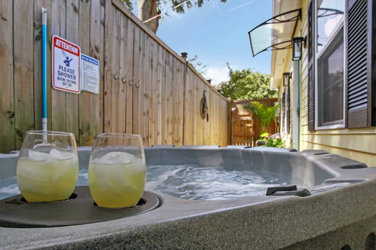 A hot tub is featured in a secluded outdoor space, with two glasses placed on the edge. The surrounding wood fence provides privacy, and a sign indicates the need for a pre-use shower. Natural light filters in, enhancing the inviting setting.