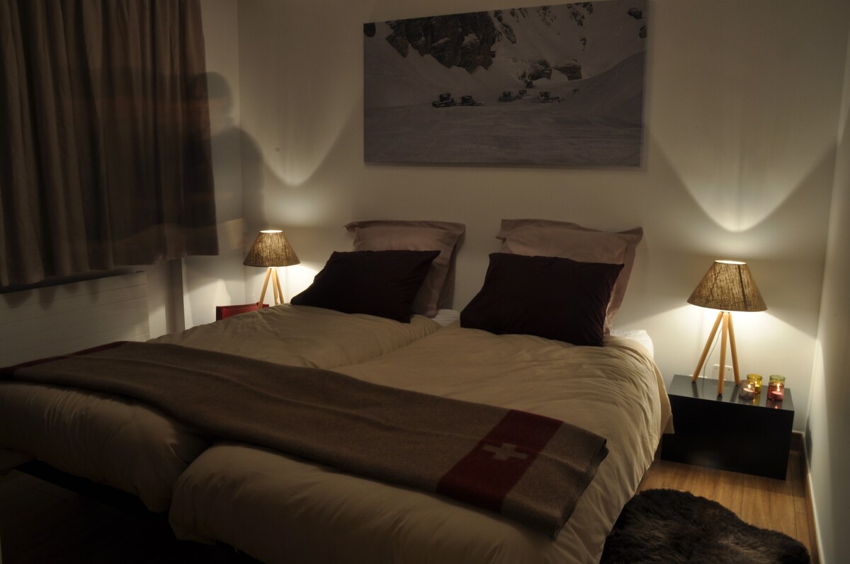 A tranquil bedroom is illuminated by soft bedside lamps, creating a calming ambiance. Two beds, dressed in neutral-coloured linens, are positioned side by side. A large wall art piece depicting a snowy landscape adds a serene touch to the space, complemented by warm hues from small decorative candles.