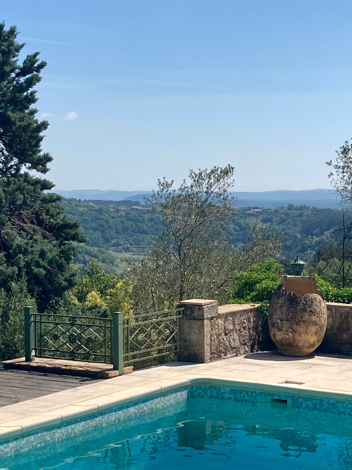 A serene outdoor space features a pool with clear water, bordered by natural stone tiles. A large decorative pot is positioned nearby, alongside lush greenery and trees. In the distance, expansive hills and a clear blue sky create a tranquil backdrop.