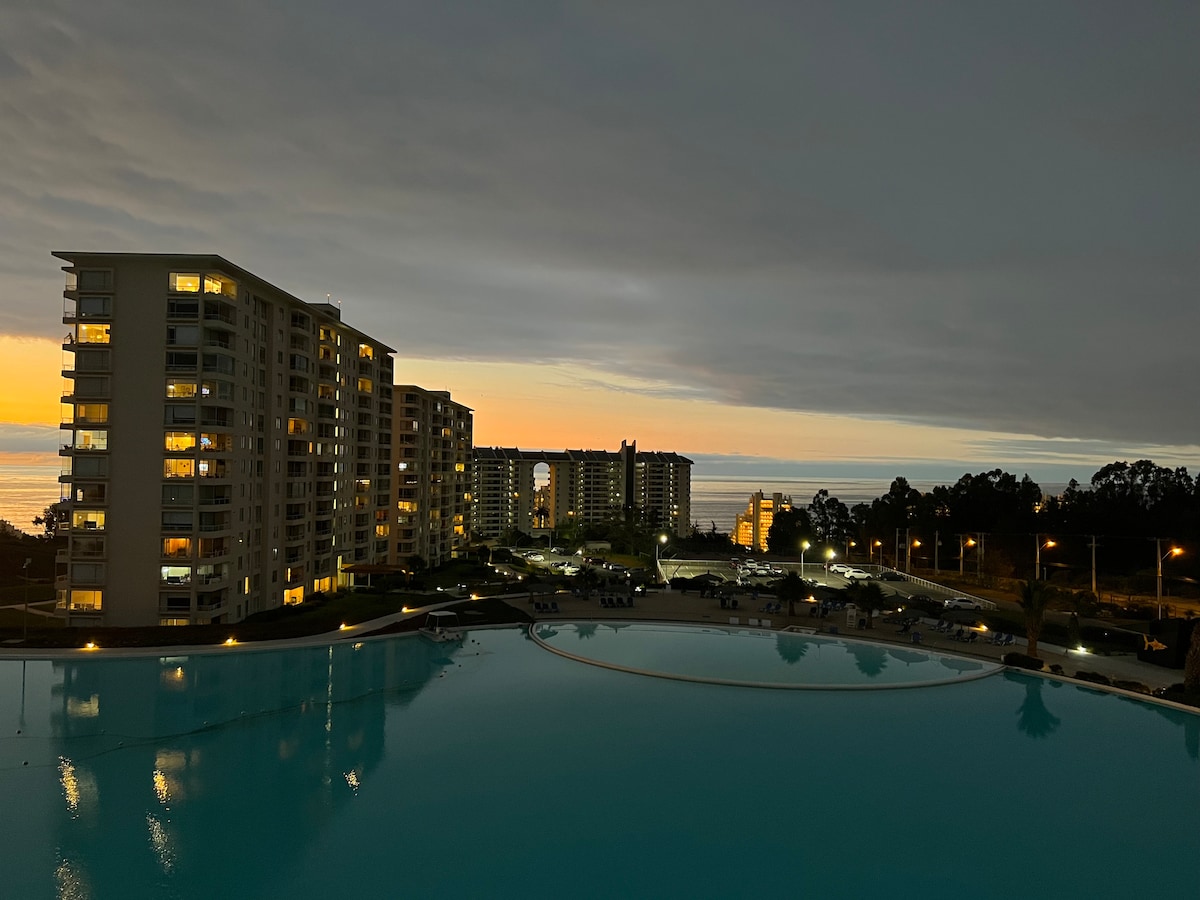 A twilight view from the balcony reveals a large swimming pool reflecting the evening sky, with several buildings lining the perimeter. The ocean is visible in the distance, while the last light of sunset casts a gentle glow over the scene.