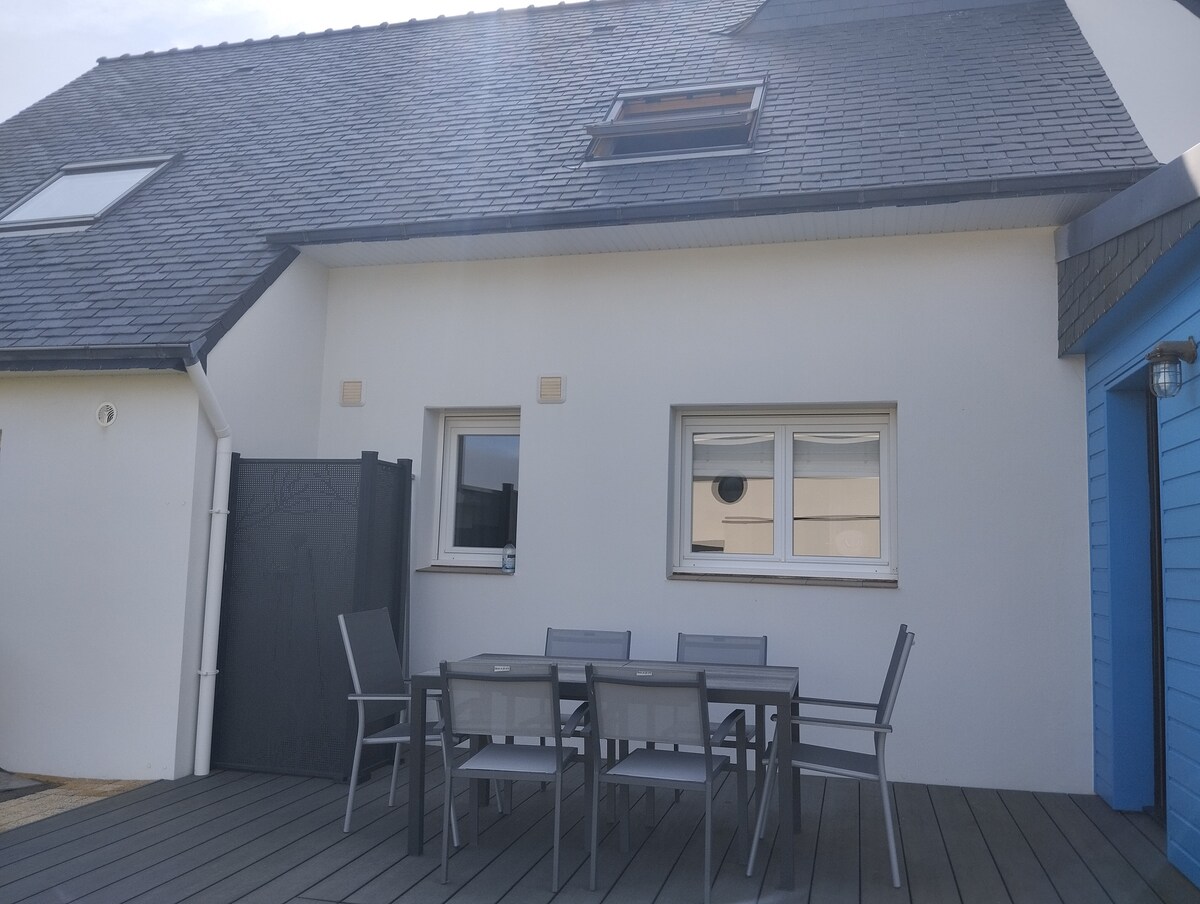 An outdoor terrace is shown, featuring a dining table surrounded by six gray chairs. The white walls of the house reflect natural light, complemented by a small blue structure nearby. A vented skylight is visible on the sloped roof.