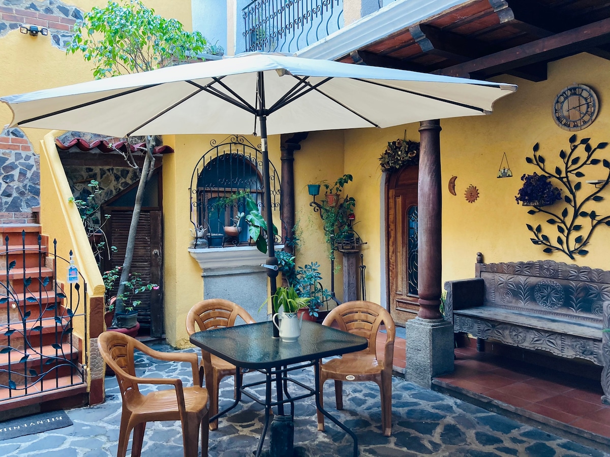 An inviting courtyard features a large umbrella over a black table surrounded by four wooden chairs. A stone path leads to a rustic bench adorned with foliage against yellow walls, while plants are elegantly arranged throughout the space, adding a touch of greenery.