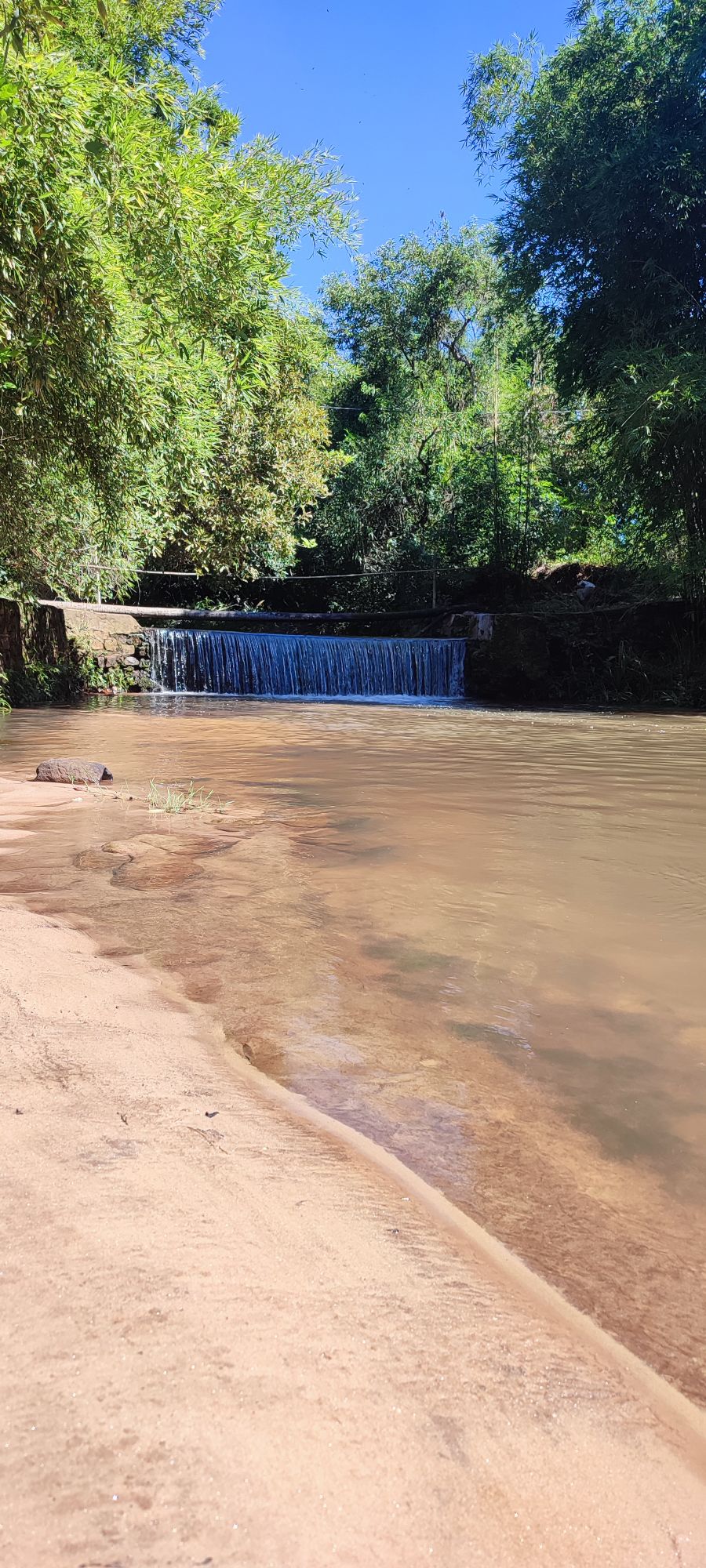 A gentle stream flows over a small waterfall, surrounded by lush greenery. Sandy shores line the water's edge, enhancing the serene natural environment. Sunlight reflects off the water's surface, creating a peaceful atmosphere.
