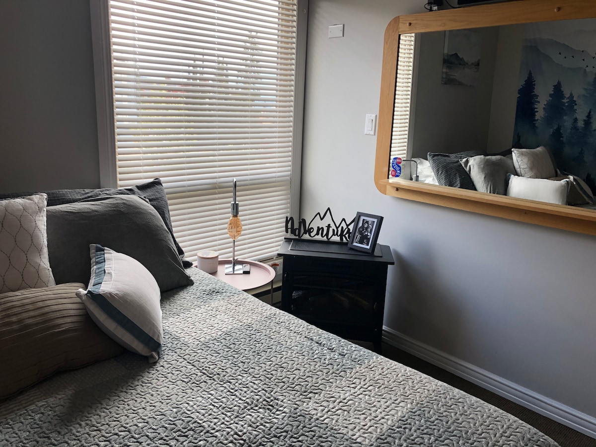 A cozy bedroom features a spacious bed adorned with textured bedding and decorative pillows. A bedside table holds a lamp and a framed photo. A large mirror reflects the inviting atmosphere, while natural light filters through the blinds of the window.