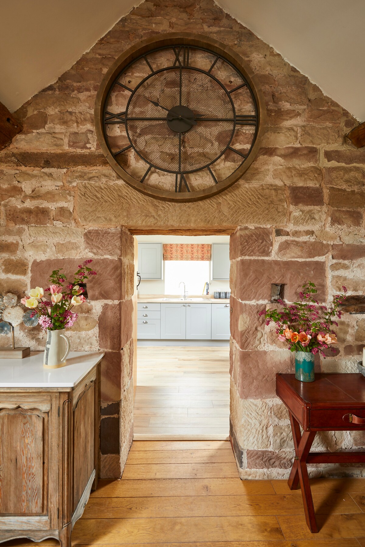 An entryway is framed by rustic stone walls, featuring a large wall clock above the archway. A wooden side table with a vase of colorful flowers sits on one side, while a chair is seen on the other, leading into a bright, inviting kitchen area.
