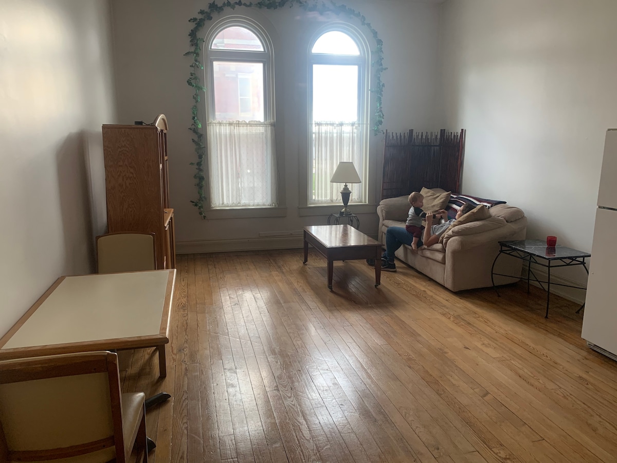 A spacious living area features large windows with arches, allowing natural light to illuminate the room. A comfortable sofa and wooden coffee table are present, along with a dining table and two chairs. Decorative vines adorn the walls, enhancing the inviting atmosphere.