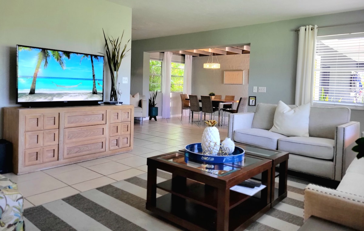 A spacious living area is presented, featuring a large flat-screen TV with a tropical scene. A comfortable sofa is positioned beside a wood coffee table with decorative items. The room benefits from natural light through the window, creating a sense of openness towards the dining area.