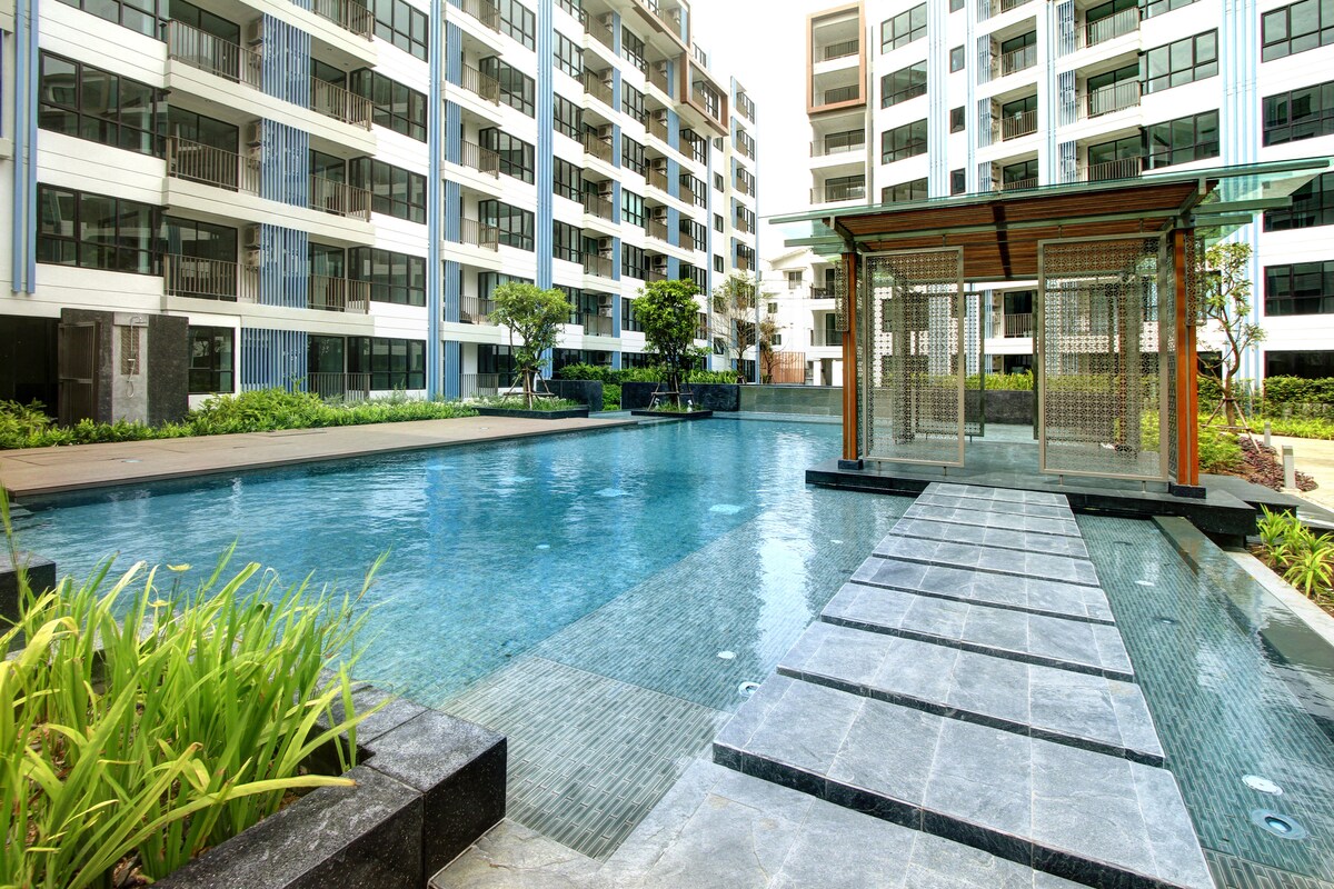 A serene swimming pool is framed by modern apartment buildings, featuring a smooth pathway of stepping stones leading across the water. Lush greenery is visible along the edges, enhancing the tranquility of the outdoor space.
