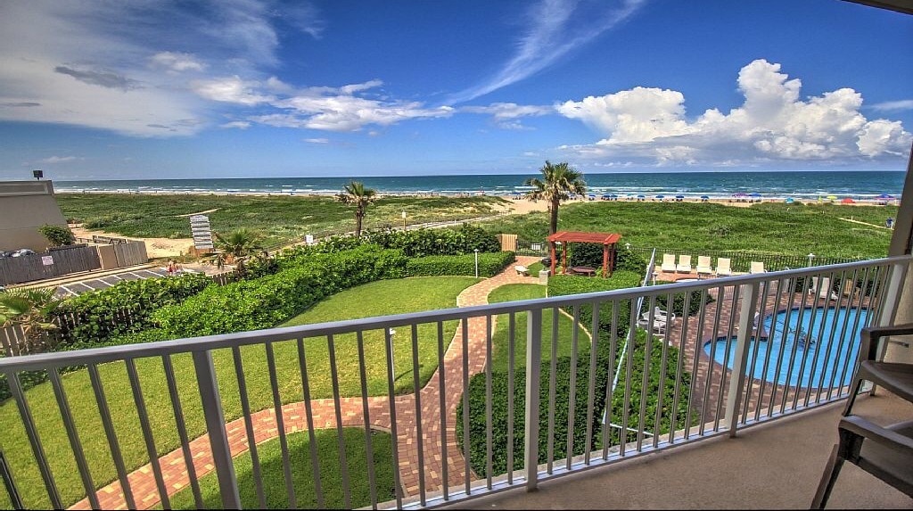 A scenic view from a balcony overlooks the beach, with green grass and a pathway leading towards the ocean. A pool area is visible, surrounded by lounge chairs. The sky features a mix of clouds and blue, creating a tranquil coastal atmosphere.