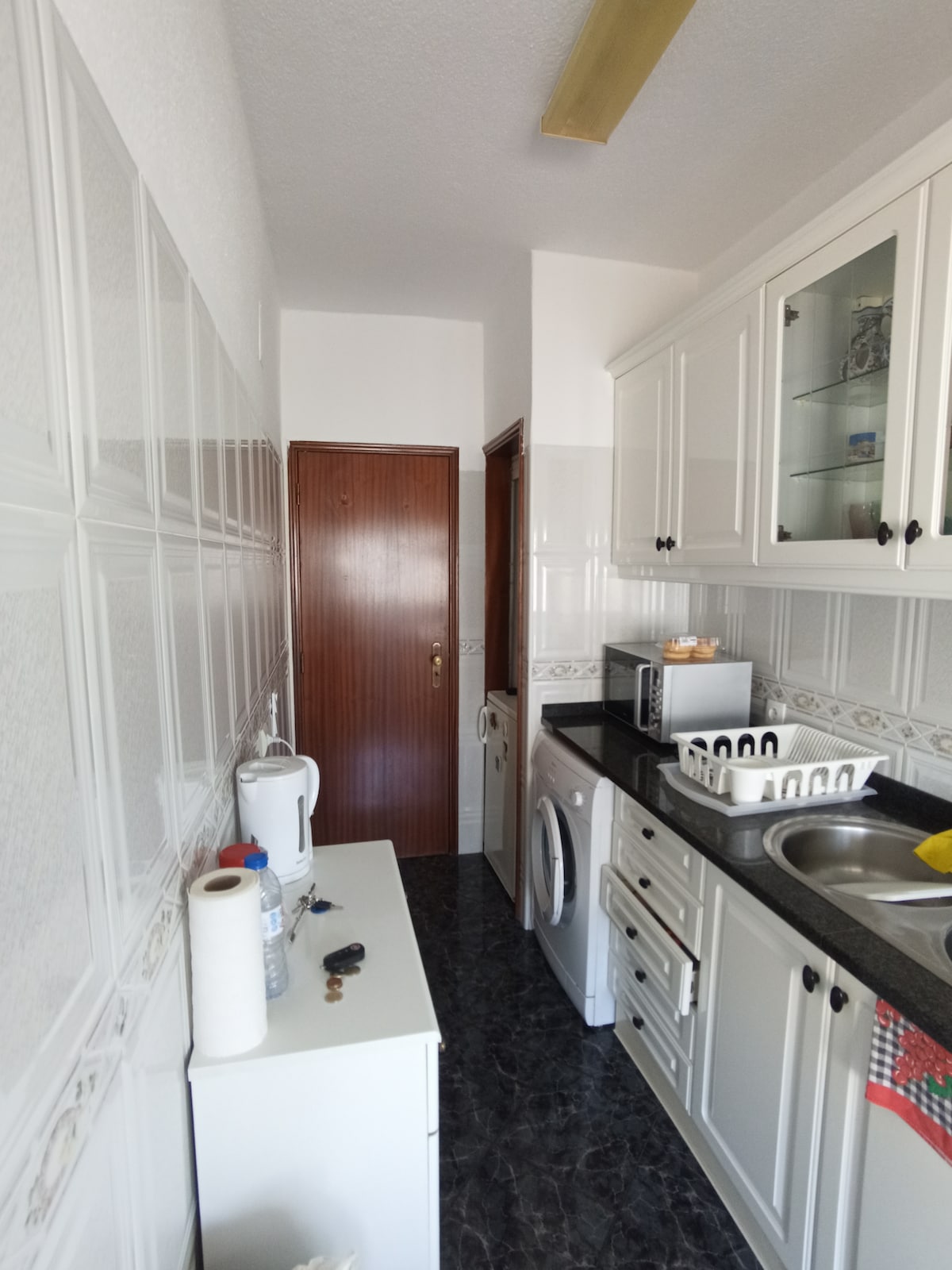 A narrow kitchen is displayed, featuring white cabinetry and dark countertop surfaces. Essential appliances, including a microwave, washing machine, and kettle, are aligned for convenience. A small white table adds functionality, while the doorframe leads to the apartment's interior.