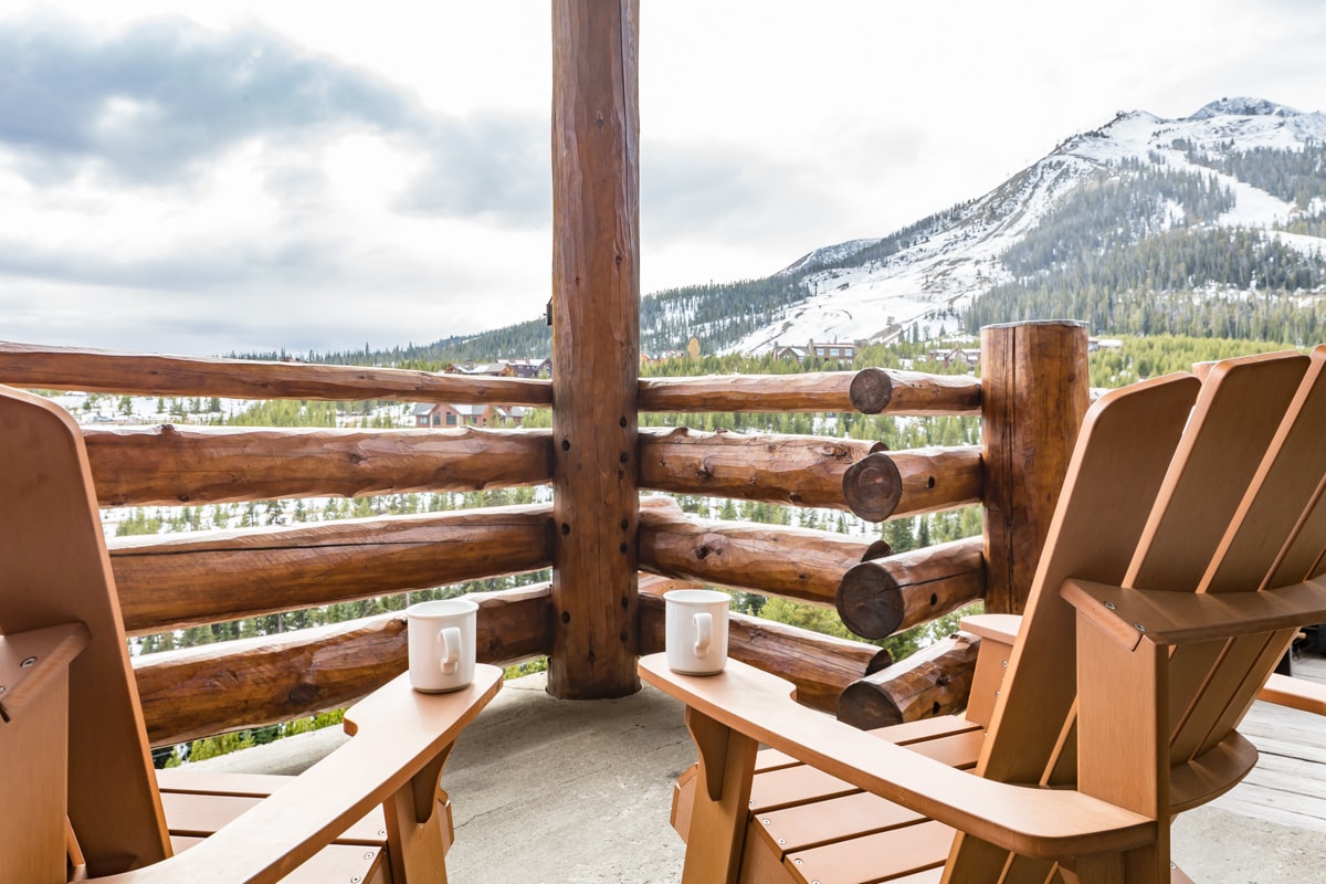 Two wooden Adirondack chairs rest on a balcony, each with a white mug placed on the surface. The view offers expansive mountain scenery with snow-capped peaks and lush green valleys, framed by the rustic log railing.