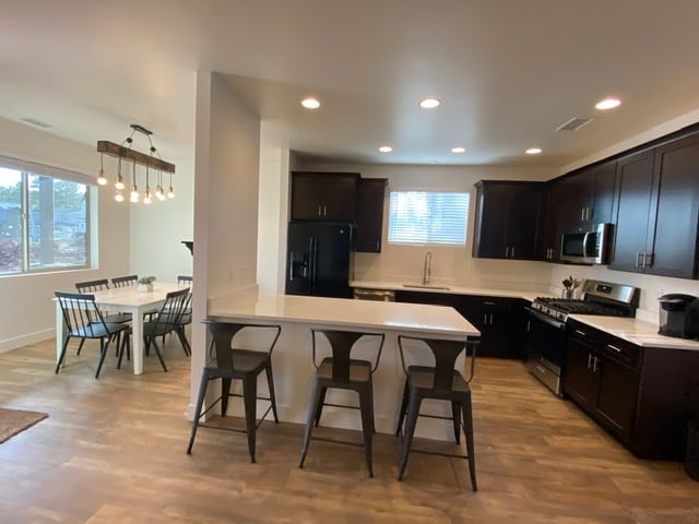 A modern kitchen features dark cabinetry and stainless-steel appliances, including a refrigerator and stove. A spacious island with seating for four is present, while a dining area with a round table and six chairs is visible in the background. Natural light is provided by a nearby window.