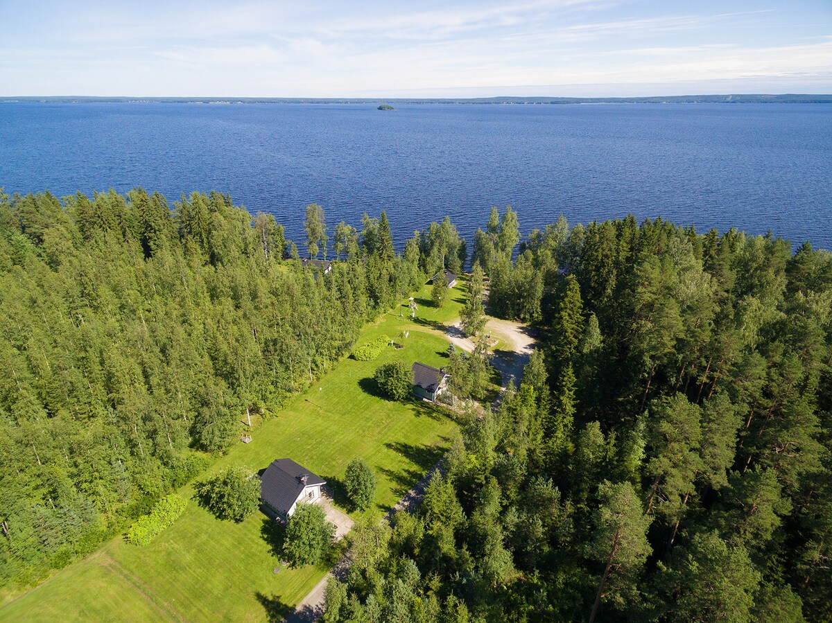 An aerial view captures the expansive, serene landscape with a calm lake bordered by dense greenery. Two cabins are nestled among well-maintained grassy areas, providing a sense of privacy and tranquility. The shoreline and distant islands are visible across the water.