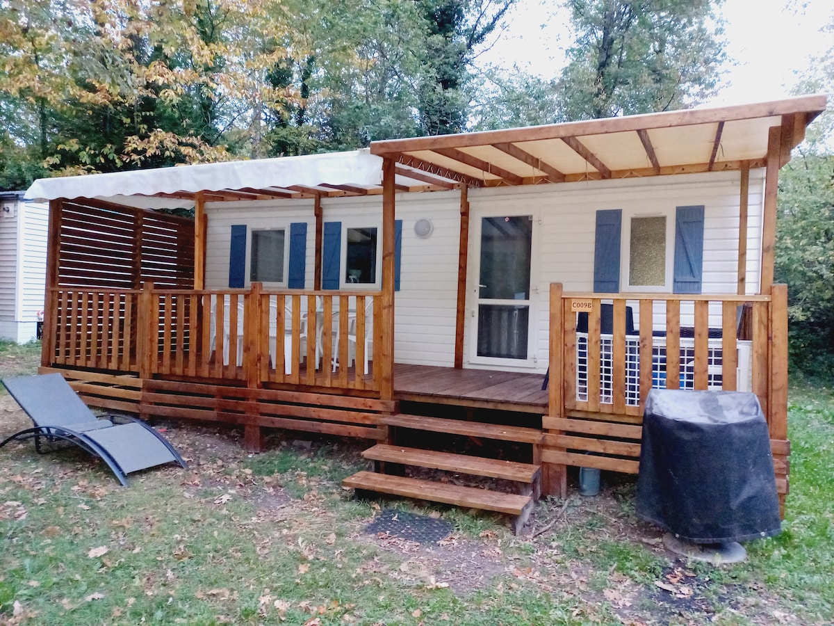 The exterior of the mobil home features a wooden deck with a pergola and a set of stairs leading to the entrance. A lounge chair is positioned nearby, and a barbecue is visible on the deck, surrounded by greenery.