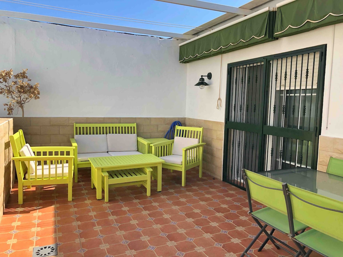 An outdoor terrace area features green patio furniture, including a seating set and dining table. The space is adorned with terracotta tiles and shaded by an awning. A door with wrought iron bars provides access to the interior, enhancing the outdoor living experience.