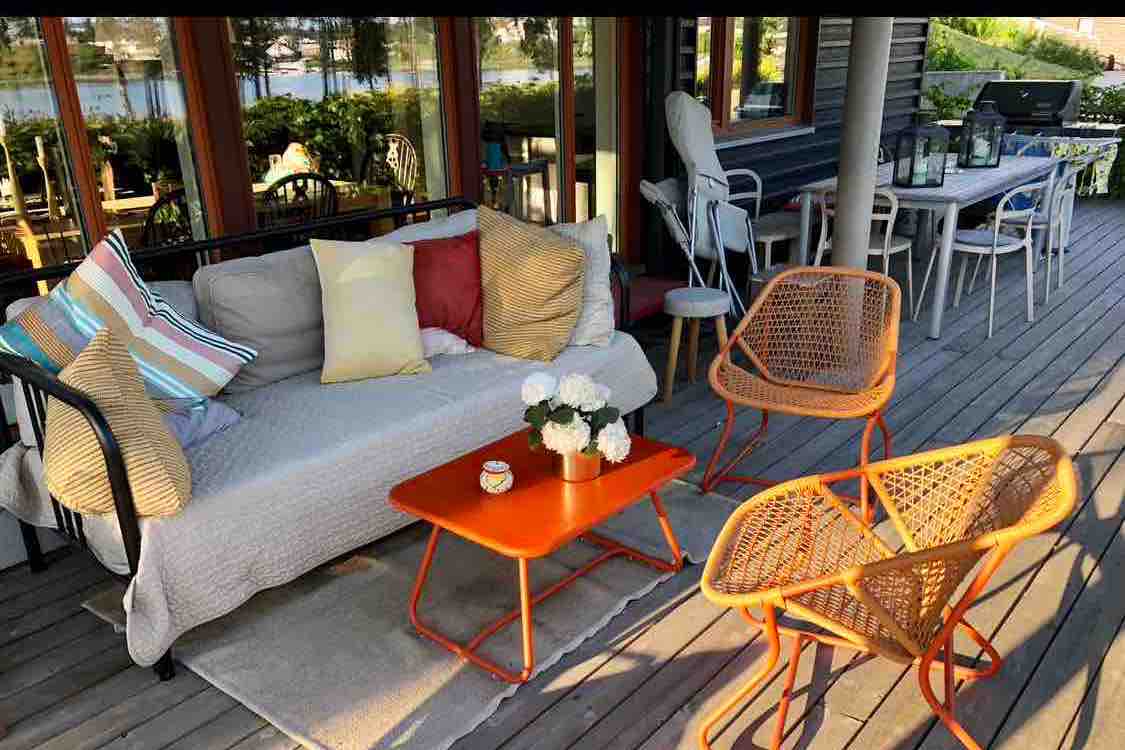 An outdoor seating area is presented with a comfortable couch adorned with decorative pillows. A bright orange coffee table sits in the center, accompanied by two rattan chairs. Natural light illuminates the space, enhancing views of greenery and water in the background.