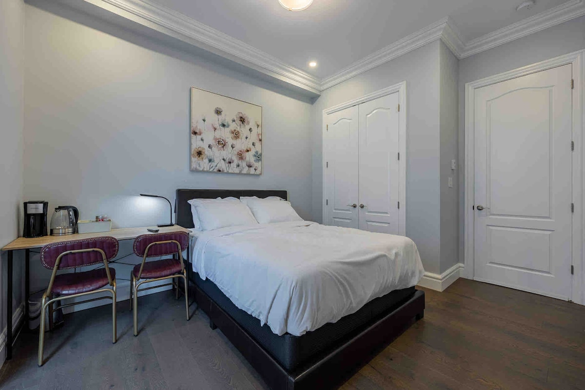 A private suite is depicted with a spacious bed and soft white linens. A small desk with two chairs is positioned to one side, alongside a coffee station. The room features light grey walls and a floral wall art piece, complemented by natural light from the nearby door.