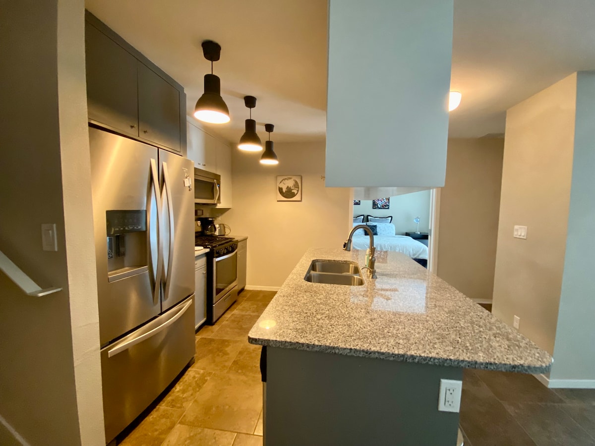 A modern kitchen features stainless steel appliances, including a large refrigerator and an oven. The polished granite countertop offers a spacious area for meal preparation. Pendant lighting hangs above the kitchen island, providing ample illumination for cooking tasks.