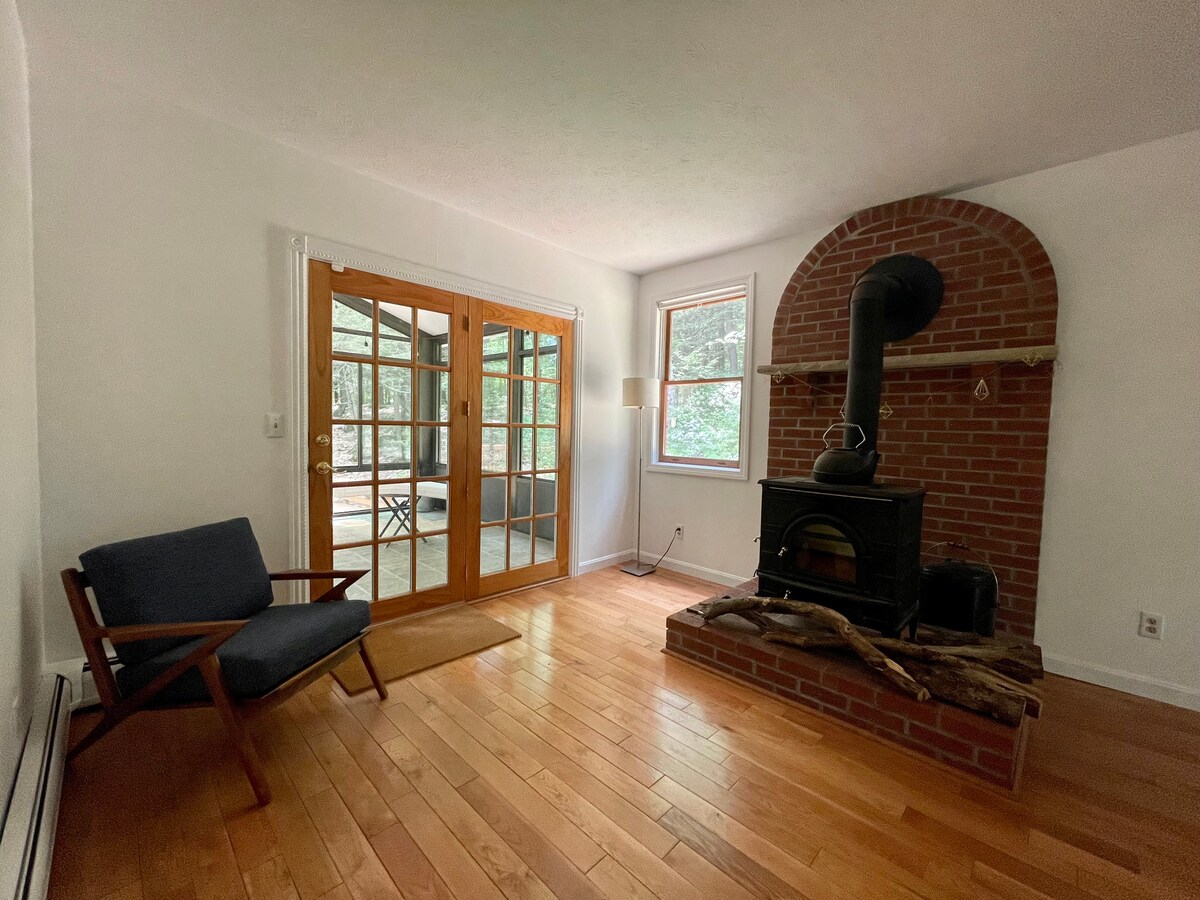 A seating area features a wooden chair positioned next to a brick fireplace with an unobtrusive black pipe. Natural light filters through a nearby window, illuminating the hardwood floor. French doors lead to a sunroom surrounded by nature.