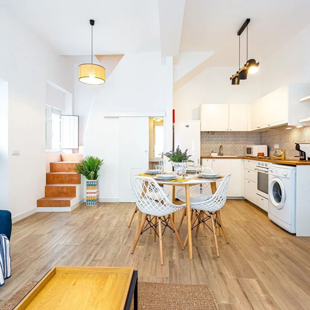 A spacious open-plan area is featured, combining a bright kitchen and dining space. A wooden dining table surrounded by four white chairs invites gatherings. Sunlight filters through windows, highlighting the modern kitchen appliances and warm wooden flooring.