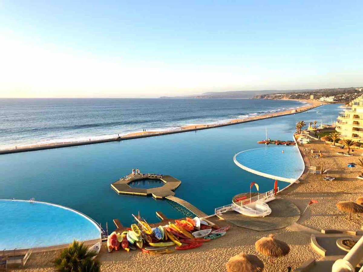 A panoramic view captures the calm ocean meeting the sky, with pools designed for relaxation lining the beachfront. Colorful kayaks and paddleboards are arranged on the sand, adding a vibrant touch to the coastal landscape. The horizon features a gentle curve leading to distant hills.