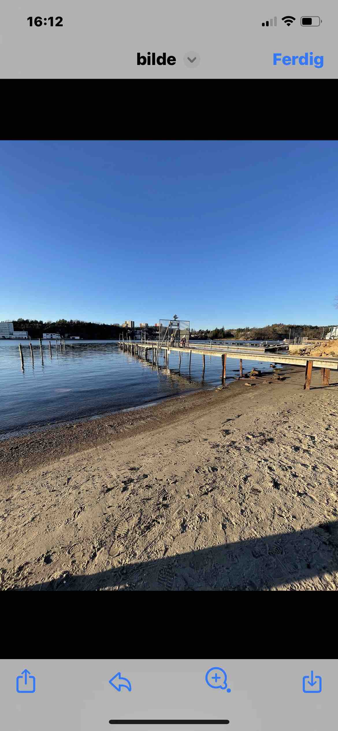 A calm shoreline features a sandy beach leading to tranquil water. A wooden pier extends into the lake, offering access to boats. The clear blue sky complements the serene atmosphere, while scenic nature is visible in the background.