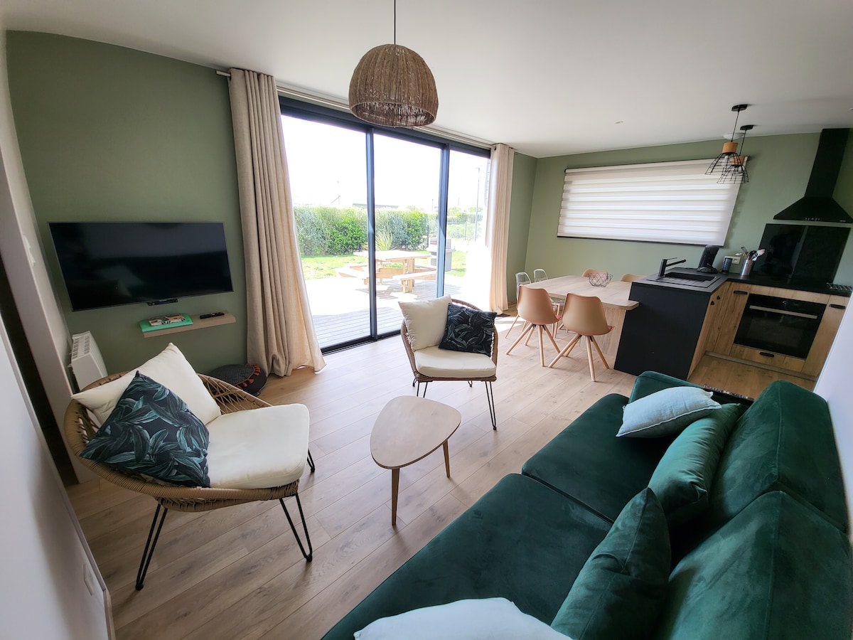 A modern living area is designed with light wood flooring and green accent walls. A comfortable dark green sofa faces a wall-mounted television. Two light-colored chairs and a dining table are visible, with sliding glass doors opening to a garden view.