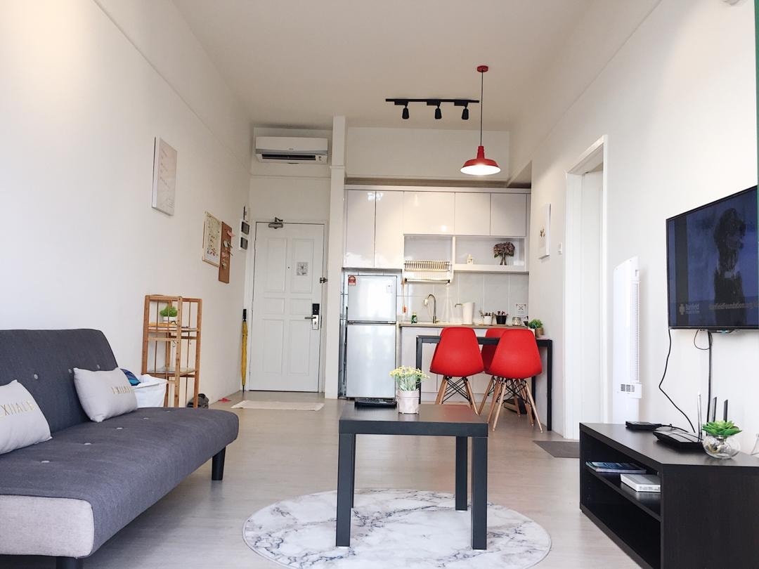A bright living area features a modern sofa bed and a marble-top coffee table. Two red chairs complement the open kitchen area, which is equipped with stainless steel appliances. A wall-mounted TV is positioned for entertainment, while air conditioning ensures comfort.