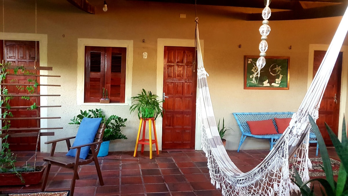An outdoor terrace features a relaxing hammock and a wooden chair. Colorful cushions add comfort to a blue furniture piece. Potted plants and a vertical garden enhance the natural ambiance, while two wooden doors lead to separate rooms, framed by earthy walls and terracotta tiles.