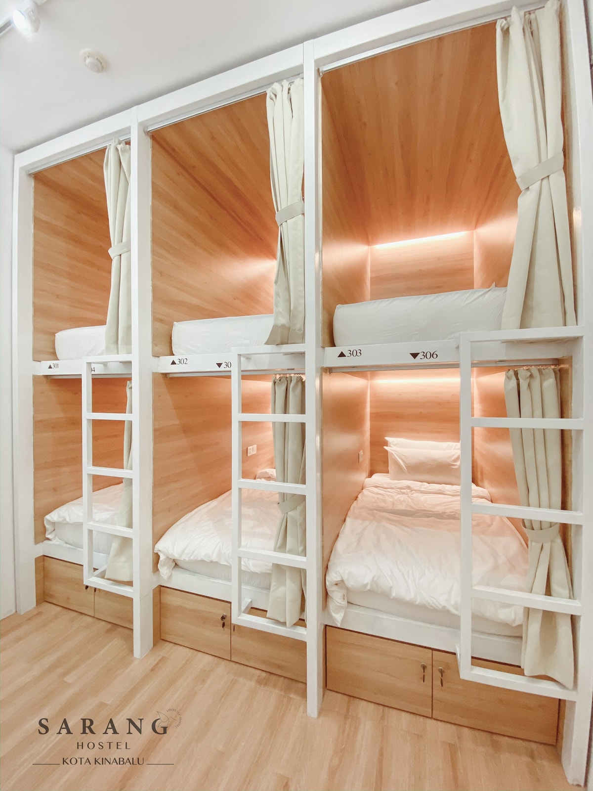 Bunk beds are arranged in a shared room, each featuring privacy curtains. Soft lighting is integrated into the wooden-paneled walls, creating a warm ambiance. The floor is polished wood, and the bedding is simple, with each bed equipped with a headboard and built-in storage underneath.