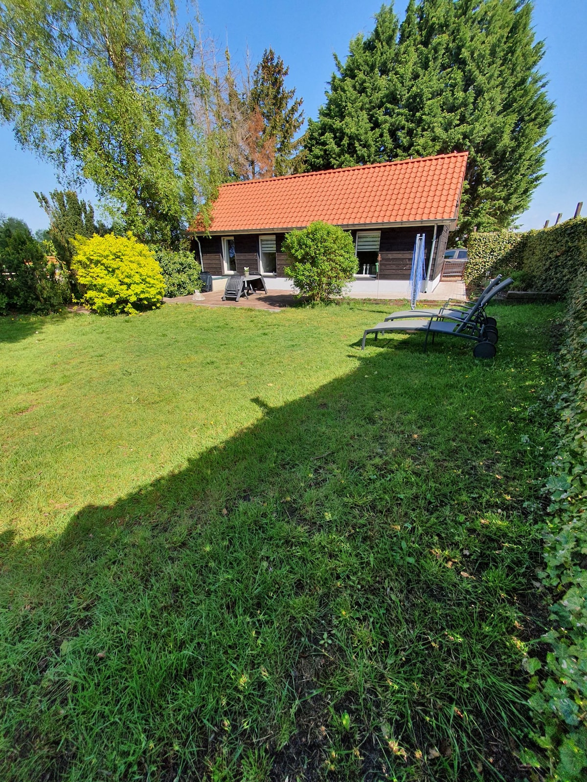 A charming guesthouse is visible amidst a well-maintained garden. The structure features a red tiled roof and large windows, framed by lush greenery. Sun loungers are positioned on the lawn, providing a cozy spot for relaxation in the open space.