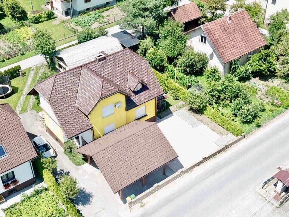 An aerial view of a yellow house with a dark brown roof is displayed. Lush green gardens surround the property, and several other homes are visible in the background. A paved driveway leads to a carport near the house, with a quiet street in front.