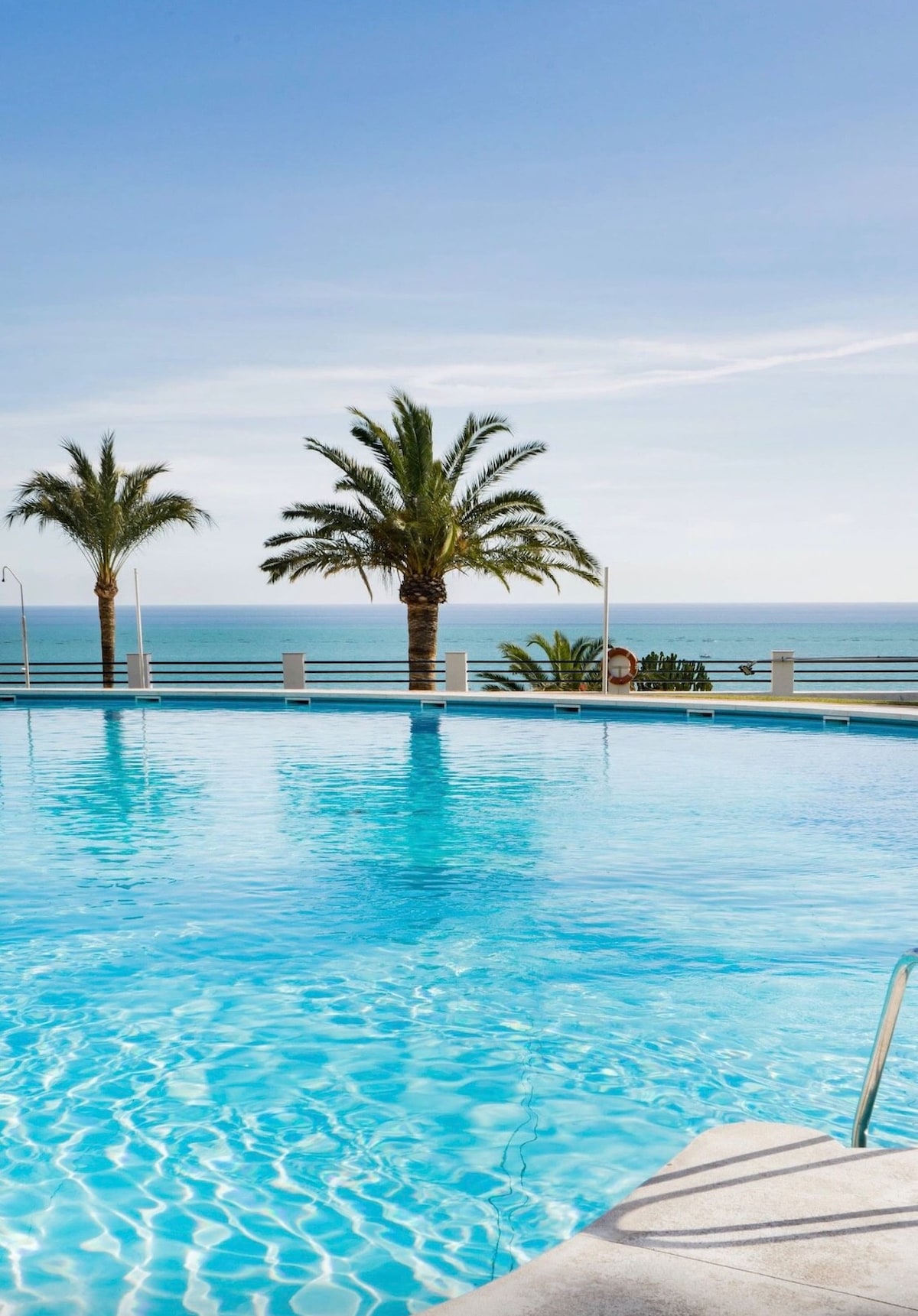 A serene swimming pool is shown surrounded by palm trees, with clear water reflecting a bright sky. Views of the sea are visible in the background, creating a calming atmosphere. The pool area is well-maintained, inviting relaxation in a sunny environment.