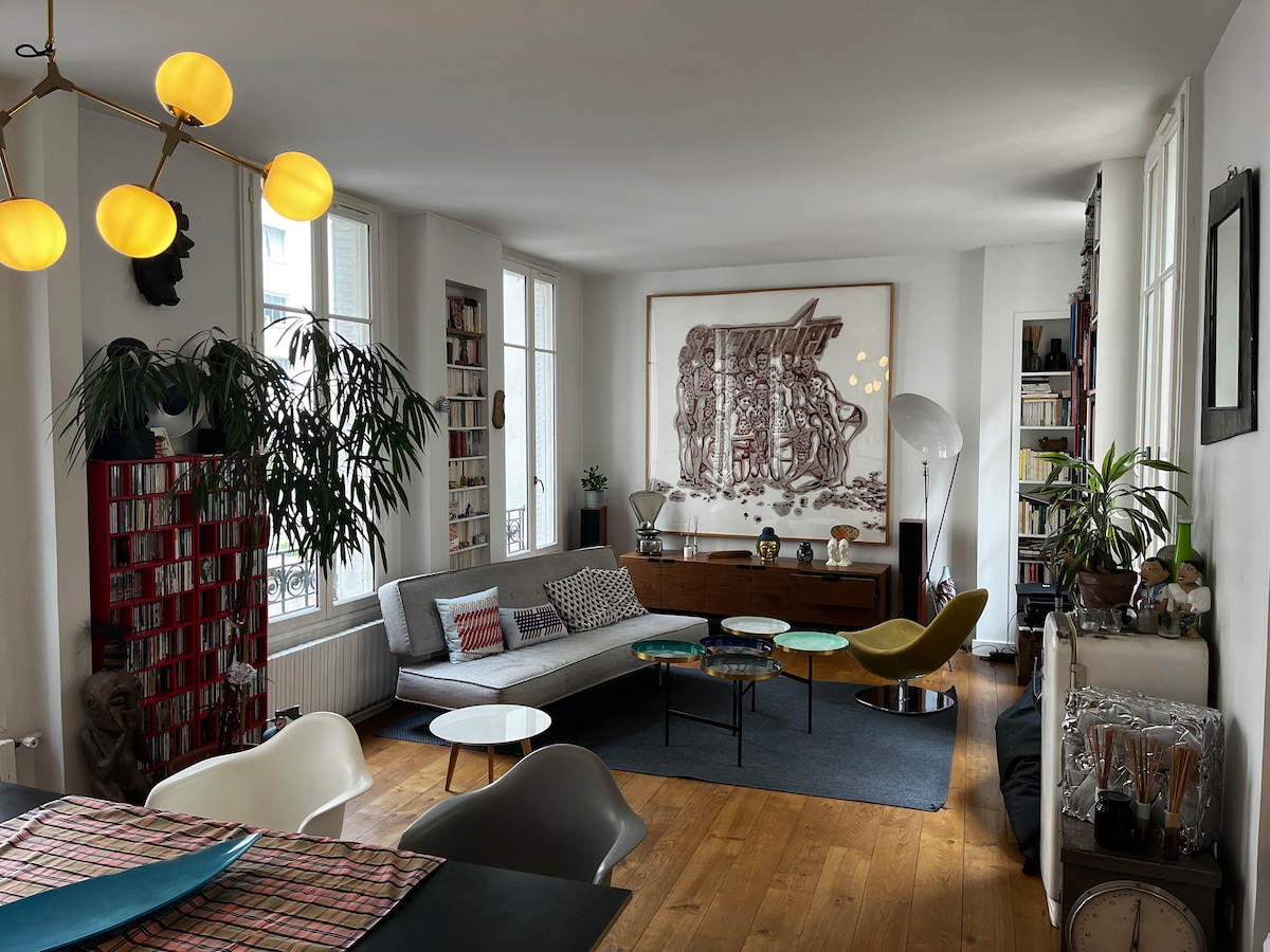 A spacious living area features large windows allowing natural light to fill the room. An abstract artwork adorns the wall behind a mid-century style wooden cabinet. A contemporary sofa, colorful accent chairs, and a dining table with a patterned tablecloth are arranged on light wooden flooring.