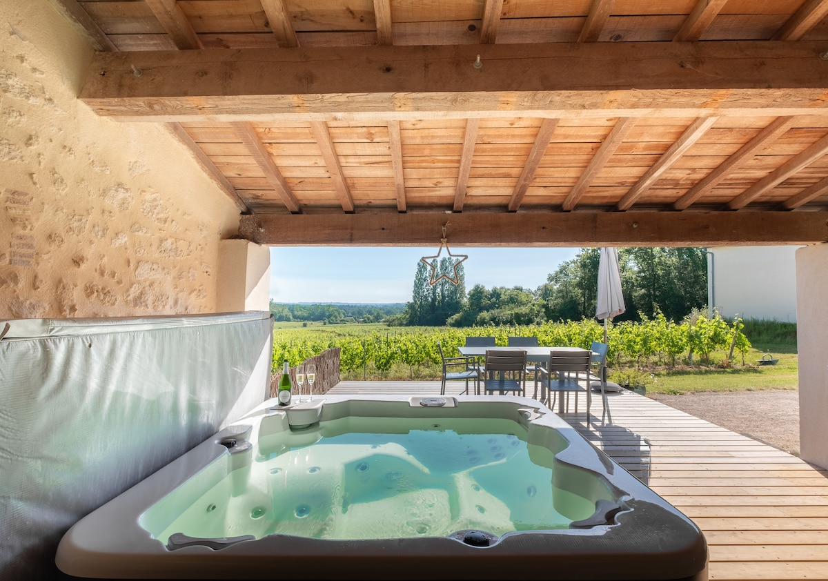 A wooden terrace provides a view of the surrounding vineyards, featuring a hot tub and outdoor dining area. A large umbrella offers shade for the seating. The rustic ceiling and open space enhance the connection with nature.