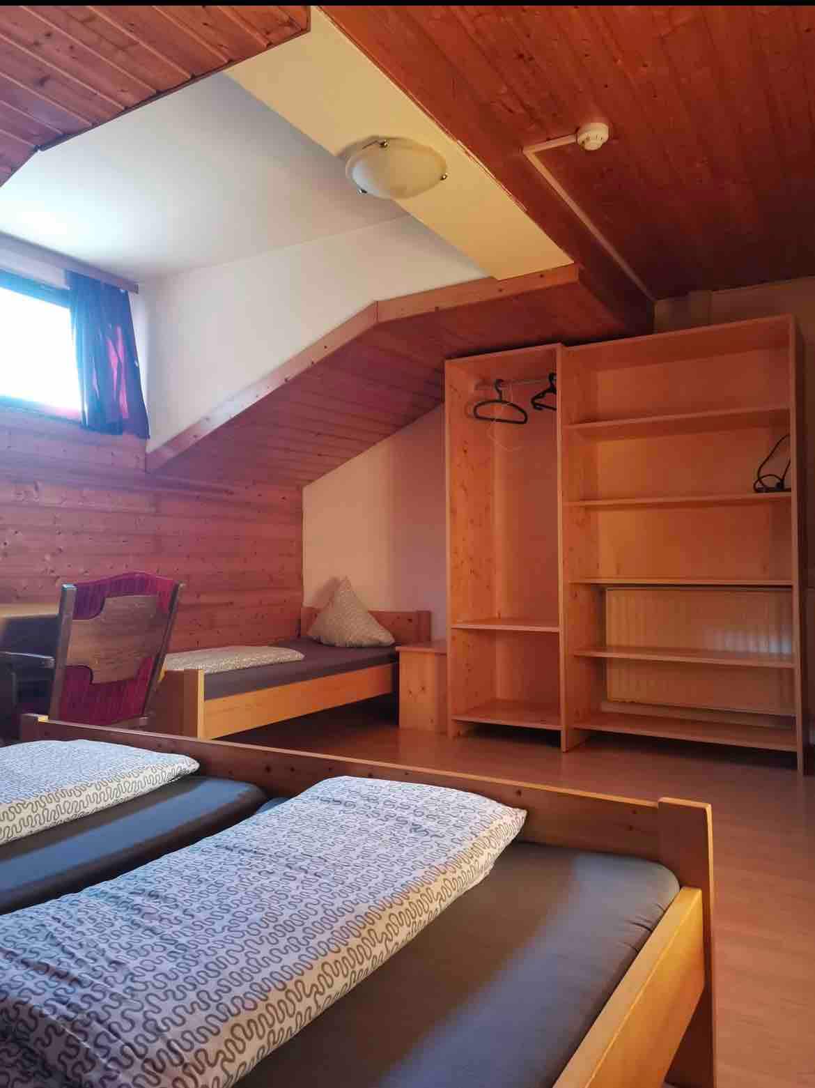 A neatly arranged room features two single beds with patterned bedding. A wooden shelving unit provides storage and additional space for personal items. A comfortable chair adds seating options, while a window allows natural light to enhance the warm wooden accents of the slanted ceiling.