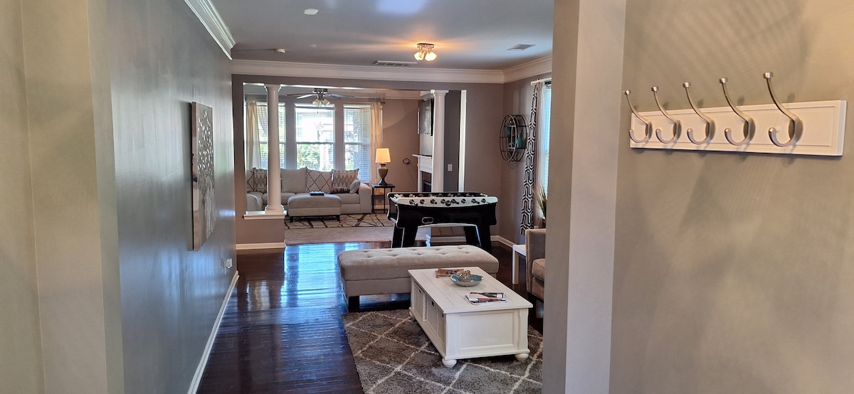 A bright and open living area is showcased, featuring large windows that invite natural light. A foosball table is positioned beside a cozy seating arrangement, which includes a coffee table and a light-colored rug. A wall-mounted coat rack is visible near the entrance.