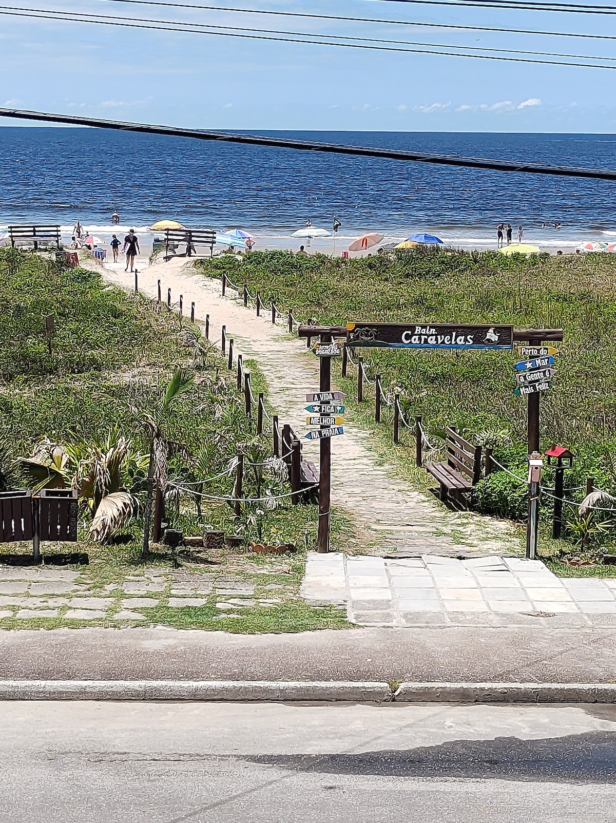 A pathway lined with grass leads to the beach, with wooden railings and signs indicating directions. In the background, beachgoers are seen enjoying the ocean, and colorful umbrellas dot the shoreline against a blue sky.