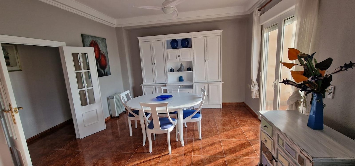 A bright dining area features a round white table surrounded by four white chairs. A white sideboard displays blue decorative items, and a small vase with flowers adds a touch of color. Light streams in through large windows, illuminating the warm-toned tiled flooring.
