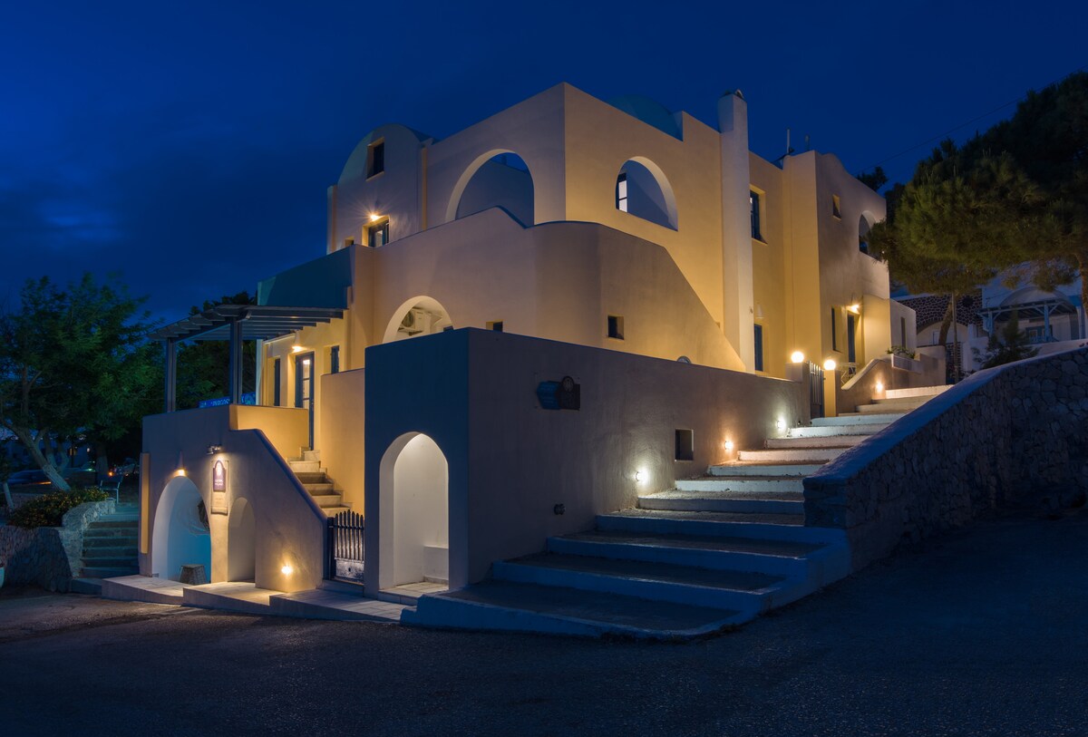 The exterior of the Cultural House is presented at night, featuring soft yellow lighting illuminating the building's Cycladic architecture. Stairs ascend towards the structure, framed by natural greenery. The design includes rounded arches and openings, highlighting the tranquil ambiance of the surroundings.