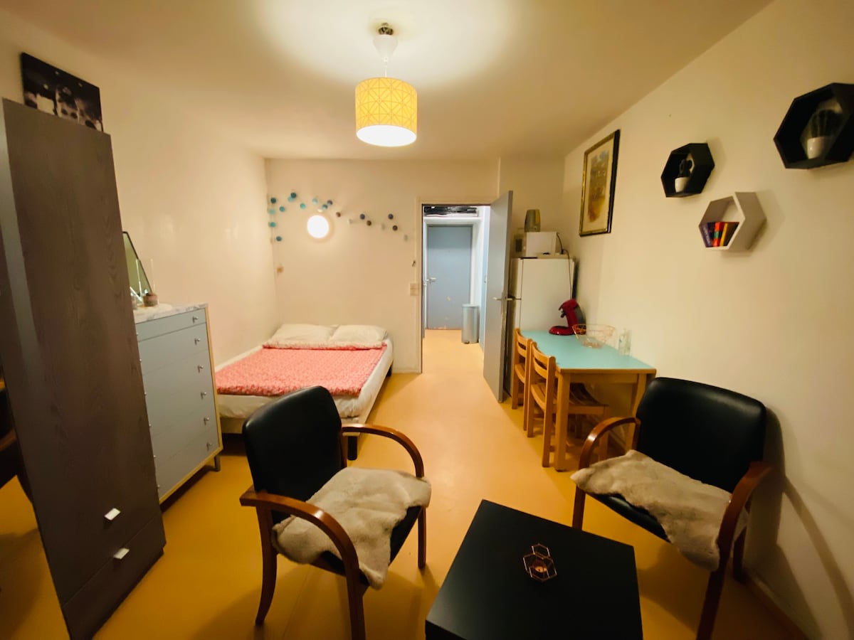 A studio space is featured, with a cozy sleeping area and a small dining table. Two black chairs are positioned around a dark table. Light-colored walls are enhanced by decorative elements, and a circular window allows natural light into the room.