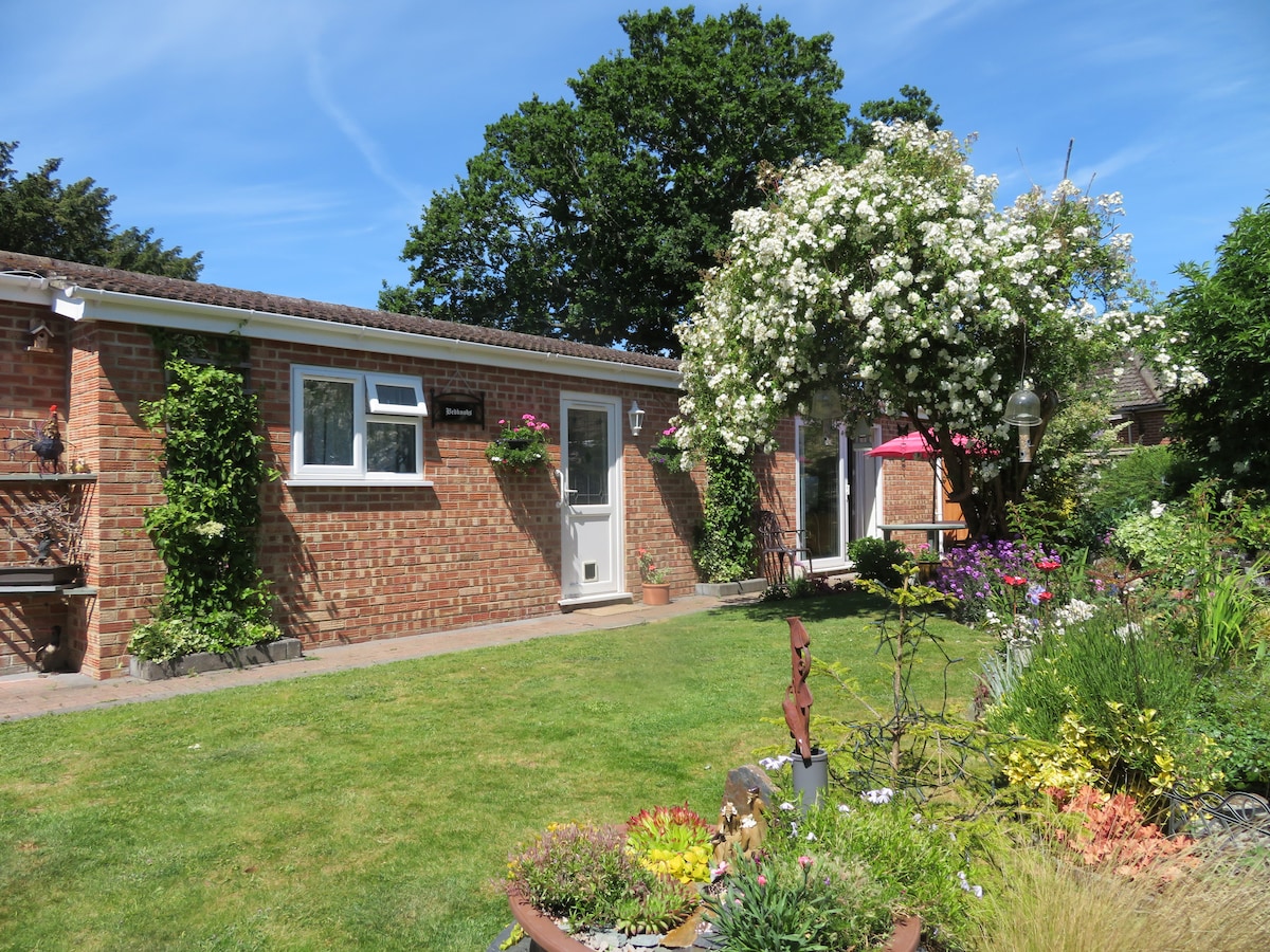 A detached self-contained property is showcased in a lush garden setting. Brightly colored flowers and greenery frame the entrance, while a door leads into the property. A flowering tree adds a touch of nature, complementing the inviting outdoor space.
