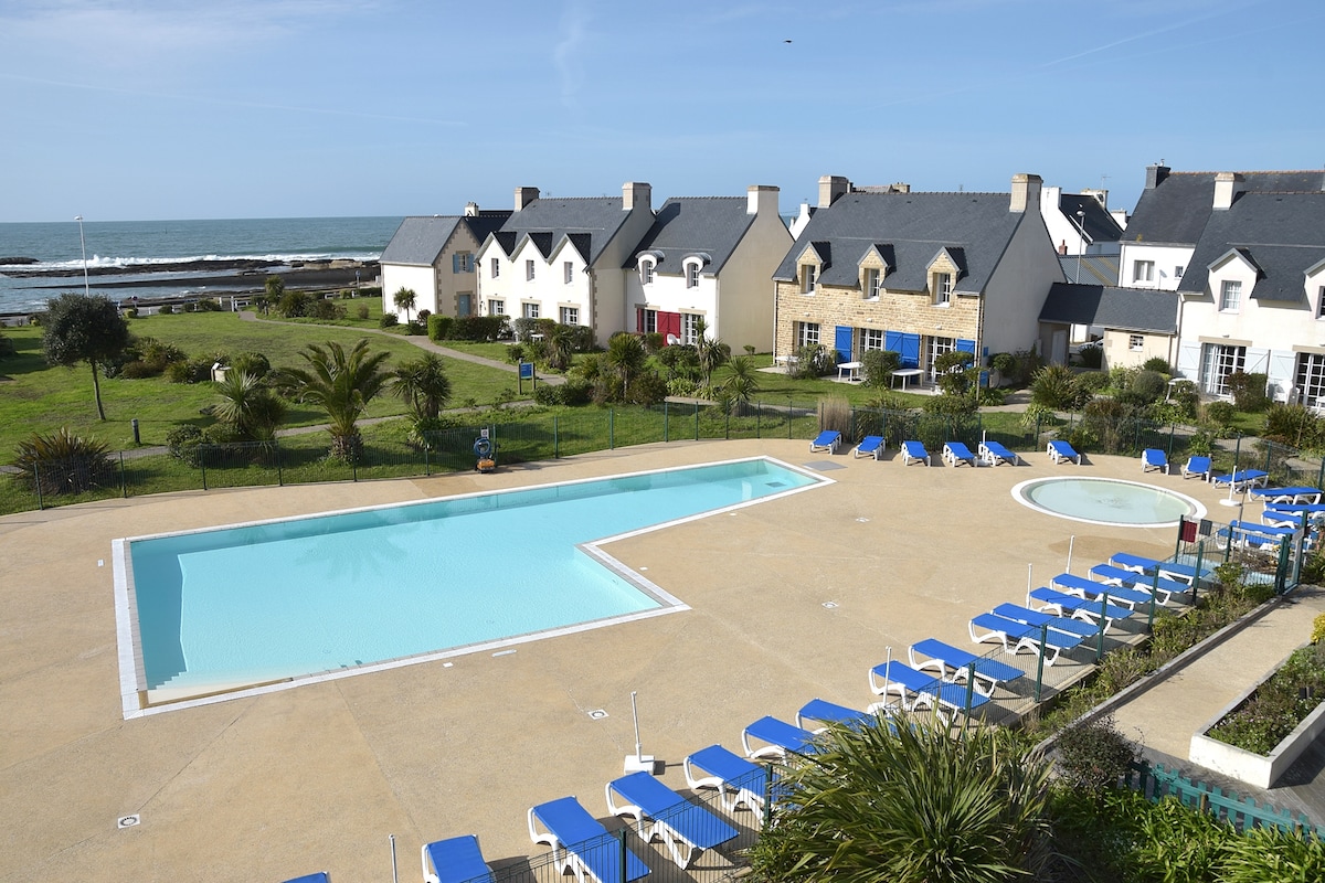 A serene outdoor area is depicted, featuring a spacious swimming pool and a smaller hot tub. Sun loungers are arranged neatly around the pool, with the ocean visible in the background. Lush greenery and well-maintained lawns frame the setting, enhancing the tranquil vacation atmosphere.