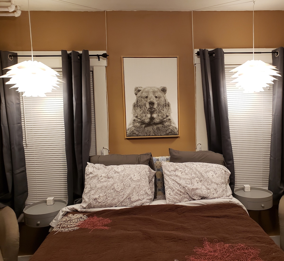 A queen bed is centered against a warm brown wall, adorned with two modern pendant lamps above. Each side of the bed features a small, round nightstand. Natural light filters through the double windows, which are dressed with dark curtains, enhancing the room's cozy ambiance.