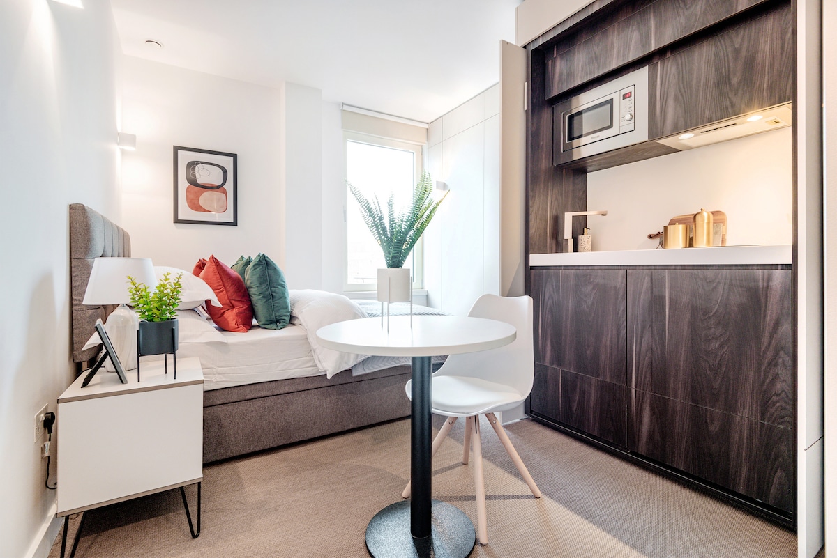 A studio apartment is displayed with a comfortable bed positioned beside a window. A round dining table is set with two white chairs, while a kitchenette is neatly integrated into cabinetry featuring a microwave and small appliances. A potted plant adds a touch of greenery.