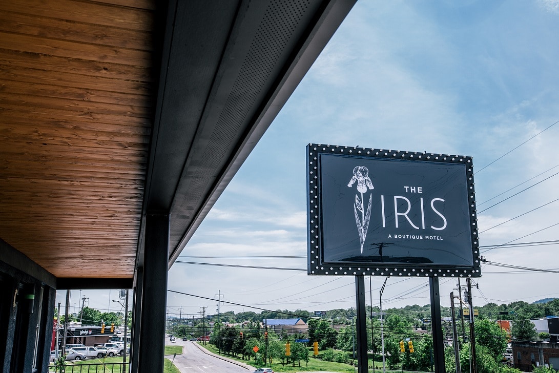 A large sign featuring 'The Iris' is prominently displayed, identifying the motel from a distance. The motel's exterior is visible with modern architectural elements and natural wood accents. A clear blue sky sets a bright backdrop, and local roads are seen in the foreground.