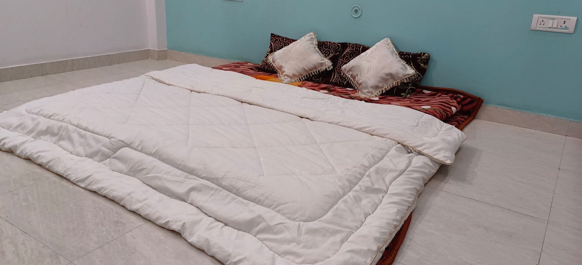 A spacious sleeping area is arranged with a large bedding quilt and decorative pillows. The flooring is tiled, contributing to a clean and open space. A calming blue wall adds a serene touch to the environment.