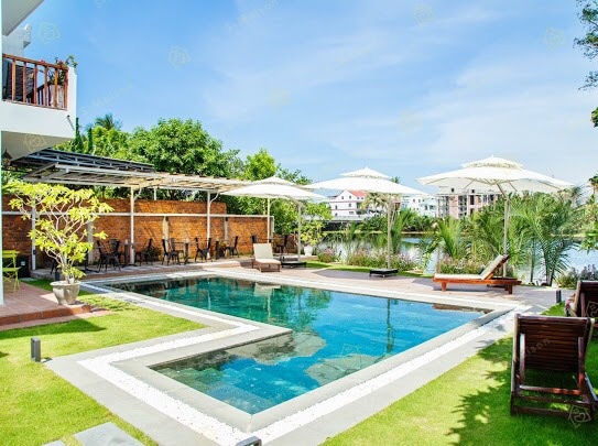 An inviting outdoor pool is surrounded by lush greenery and sun loungers. Umbrellas provide shade for relaxation. The tranquil water reflects the blue sky, creating a serene atmosphere perfect for unwinding.