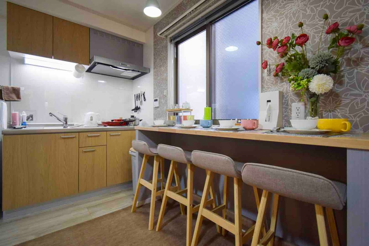 The kitchen area features warm wooden cabinets and a modern stove. A countertop with four stools is visible. Natural light enters through a window adorned with floral decor, illuminating details like a kettle, cups, and fresh flowers on the counter.